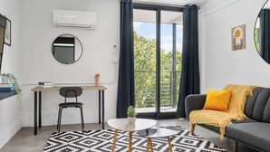 Una sala de estar moderna con un sofá, una mesa de centro, una silla y una mesita con una planta. Se observa un ventanal con cortinas, un equipo de aire acondicionado y un espejo montado en la pared.