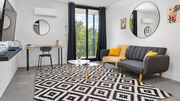 Una sala de estar moderna con un sofá gris, alfombra con estampado blanco y negro, y un comedor con una mesa pequeña y sillas.