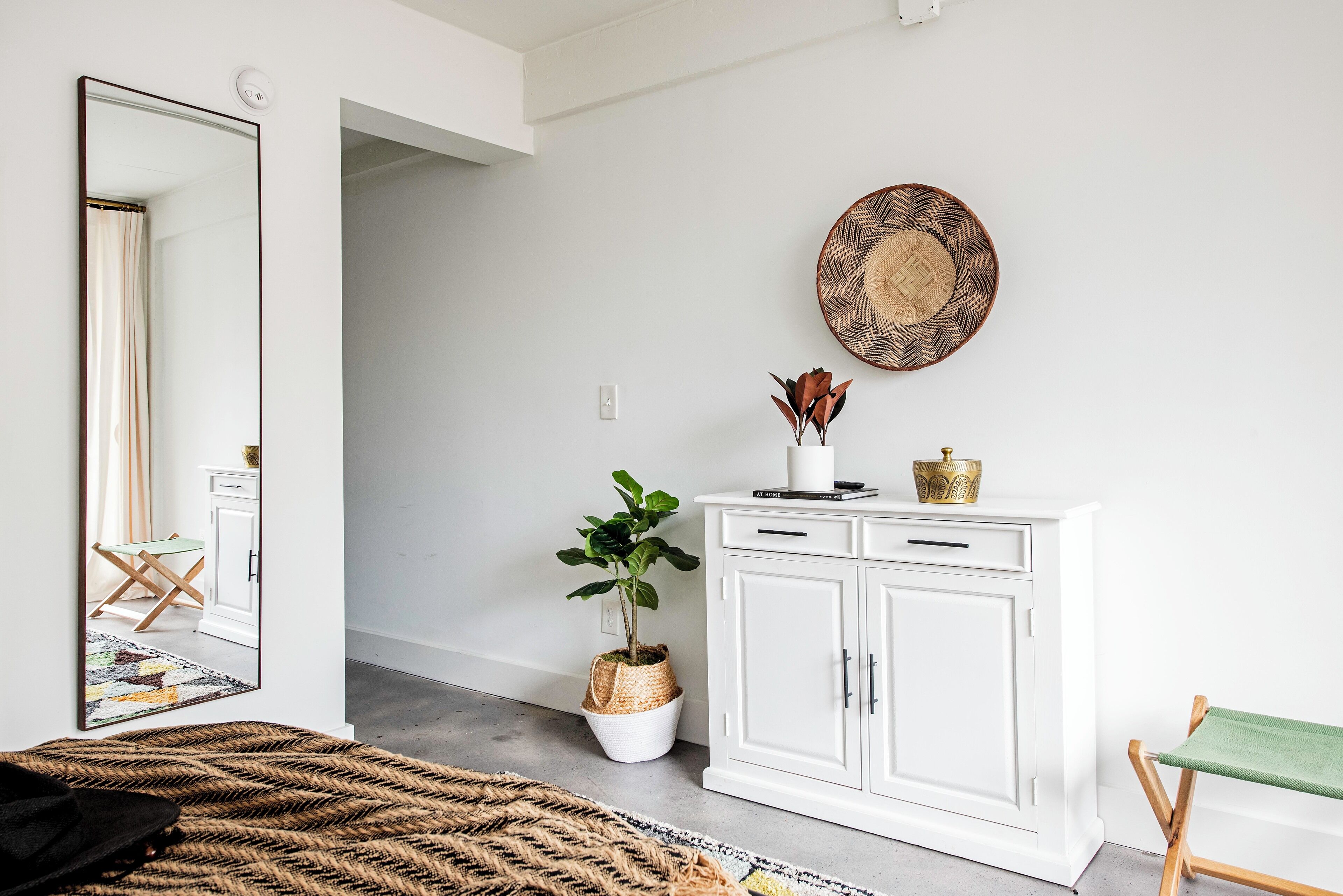 Una habitación con un espejo grande, un armario blanco, una cesta de mimbre con una planta y una alfombra tejida.