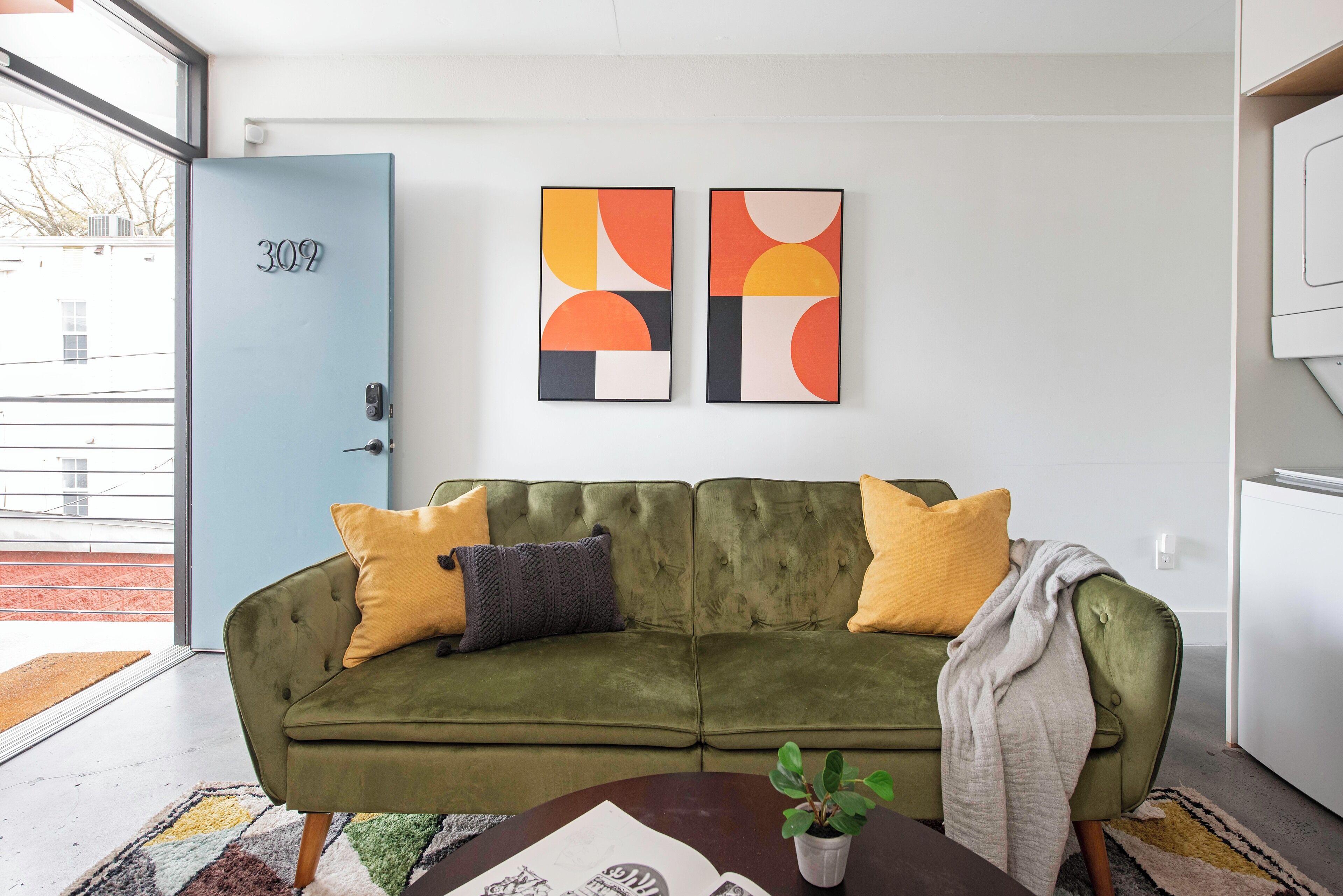 A green sofa with yellow and gray cushions, a small table with a plant, and abstract art on the wall.
