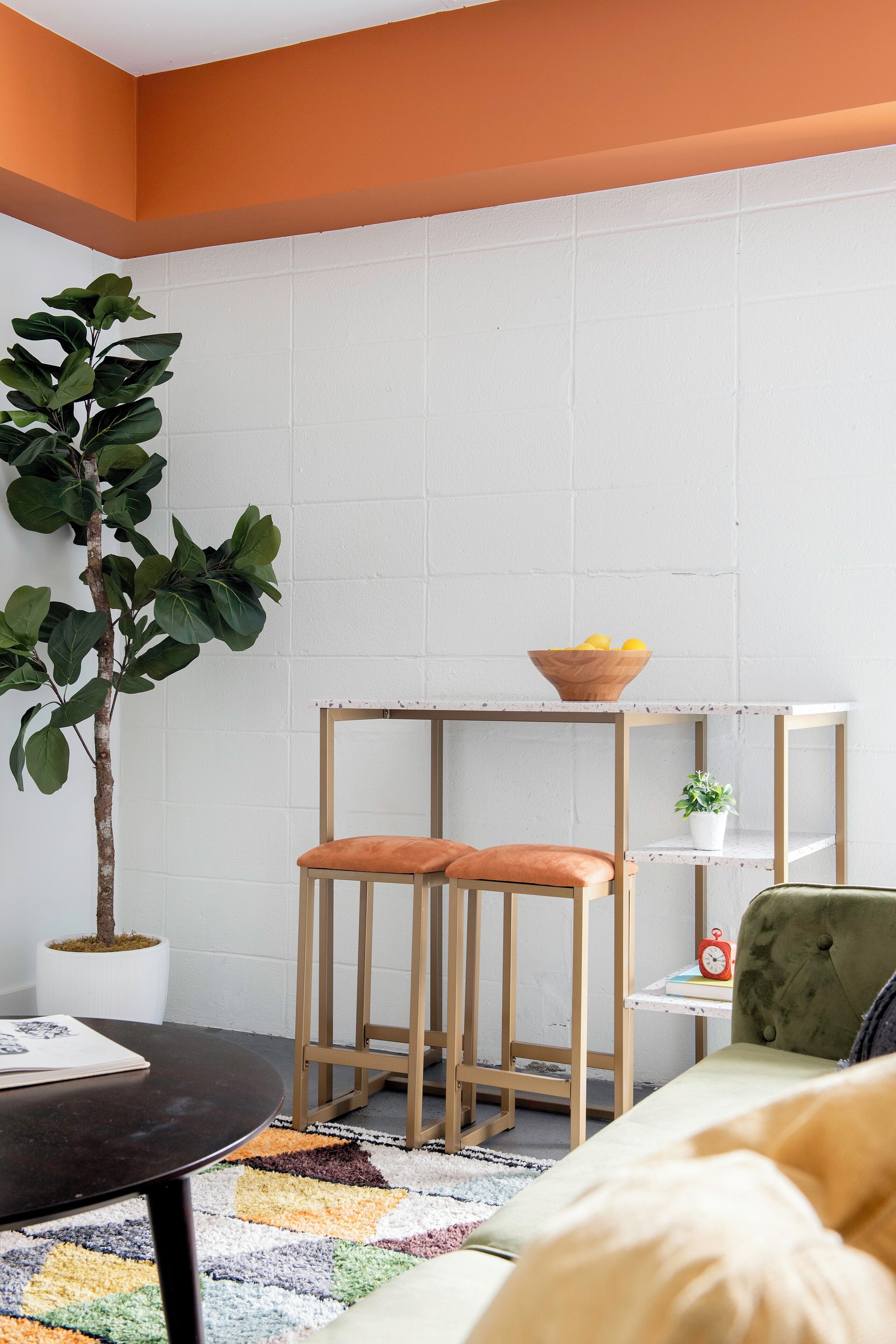 A room with a white brick wall, a wooden chair with orange cushions, a small wooden table with a bowl of lemons, a potted plant, and a green armchair.