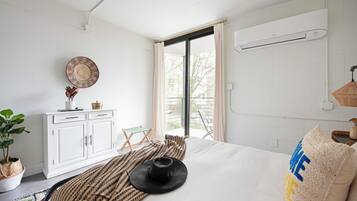 A bedroom with a bed, a white wardrobe, a small table with a plant, a window with curtains, and a balcony view.
