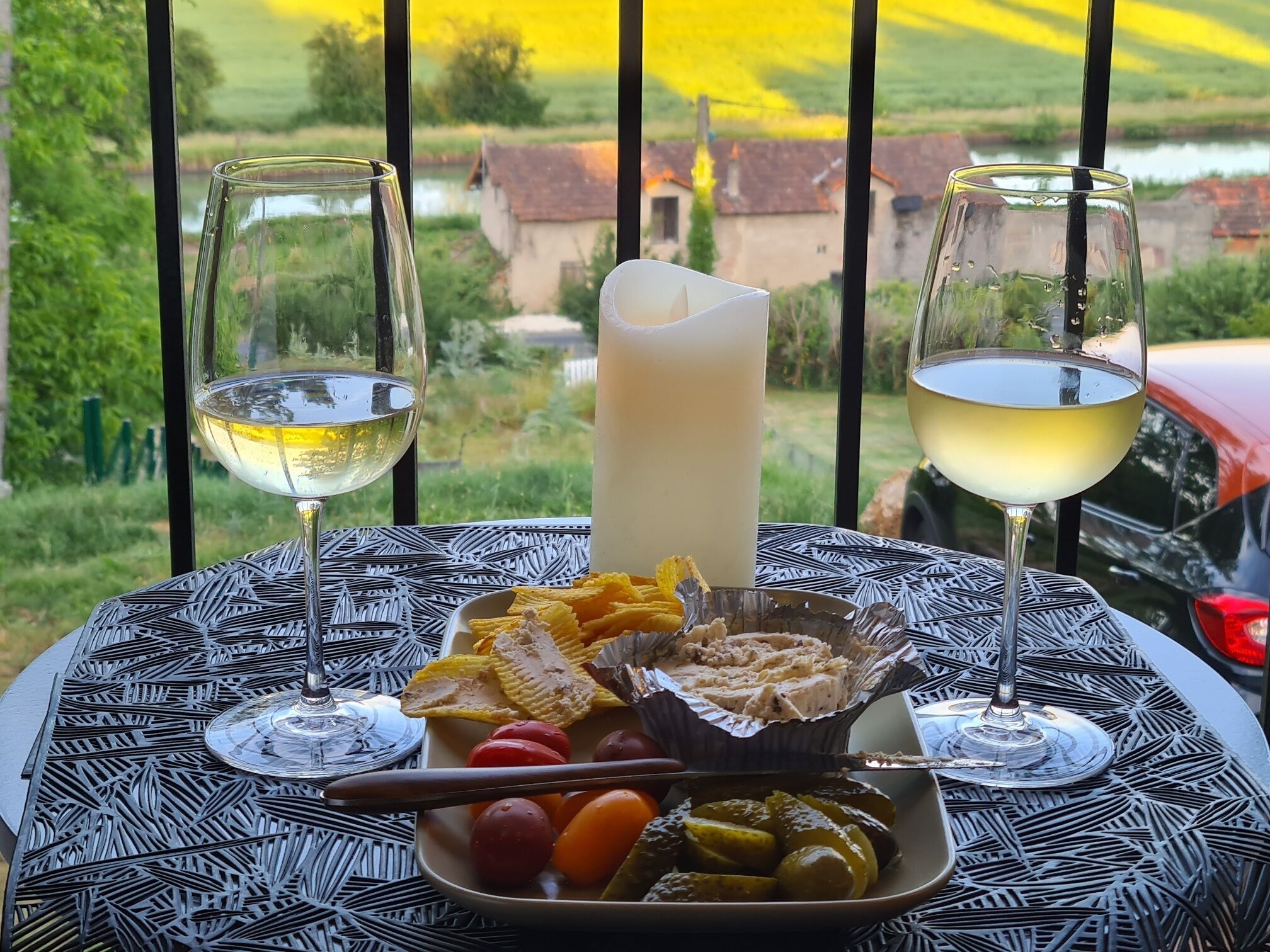 Una mesa con dos copas de vino blanco, una vela y un plato de comida que incluye pepinillos, tomates y galletas.