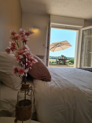 Une chambre avec un lit, un vase rempli de fleurs et une vue sur une table et un parasol situés à l’extérieur.