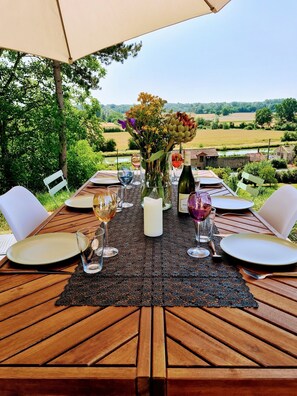 Une table à manger installée à l’extérieur, offrant une vue sur un paysage rural, et mise avec des assiettes, des verres et un vase rempli de fleurs.