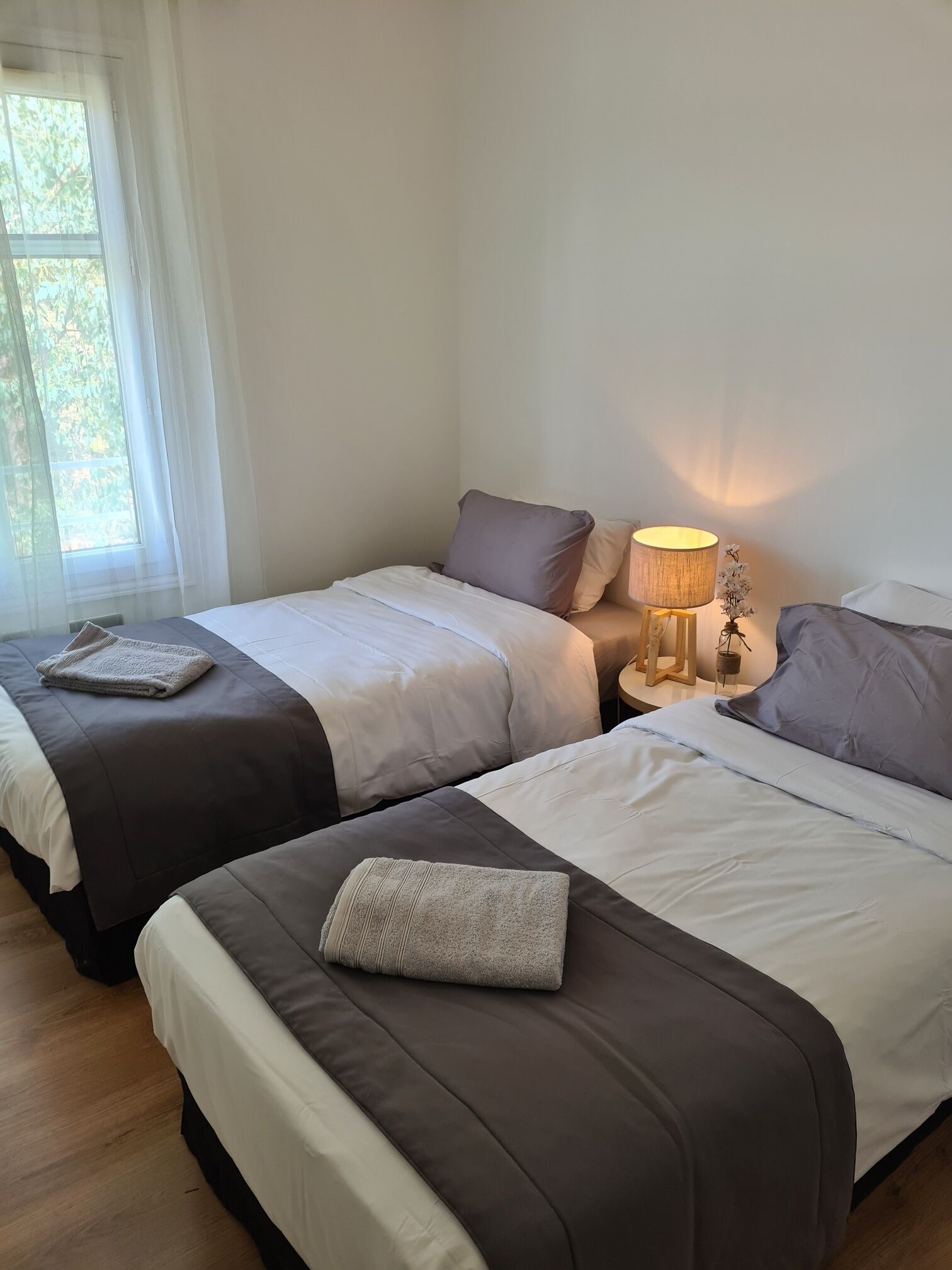 A hotel room with two beds, a bedside table with a lamp, and a window with sheer curtains.