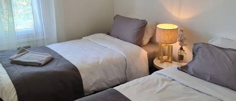 A hotel room with two beds, a bedside table with a lamp, and a window with sheer curtains.