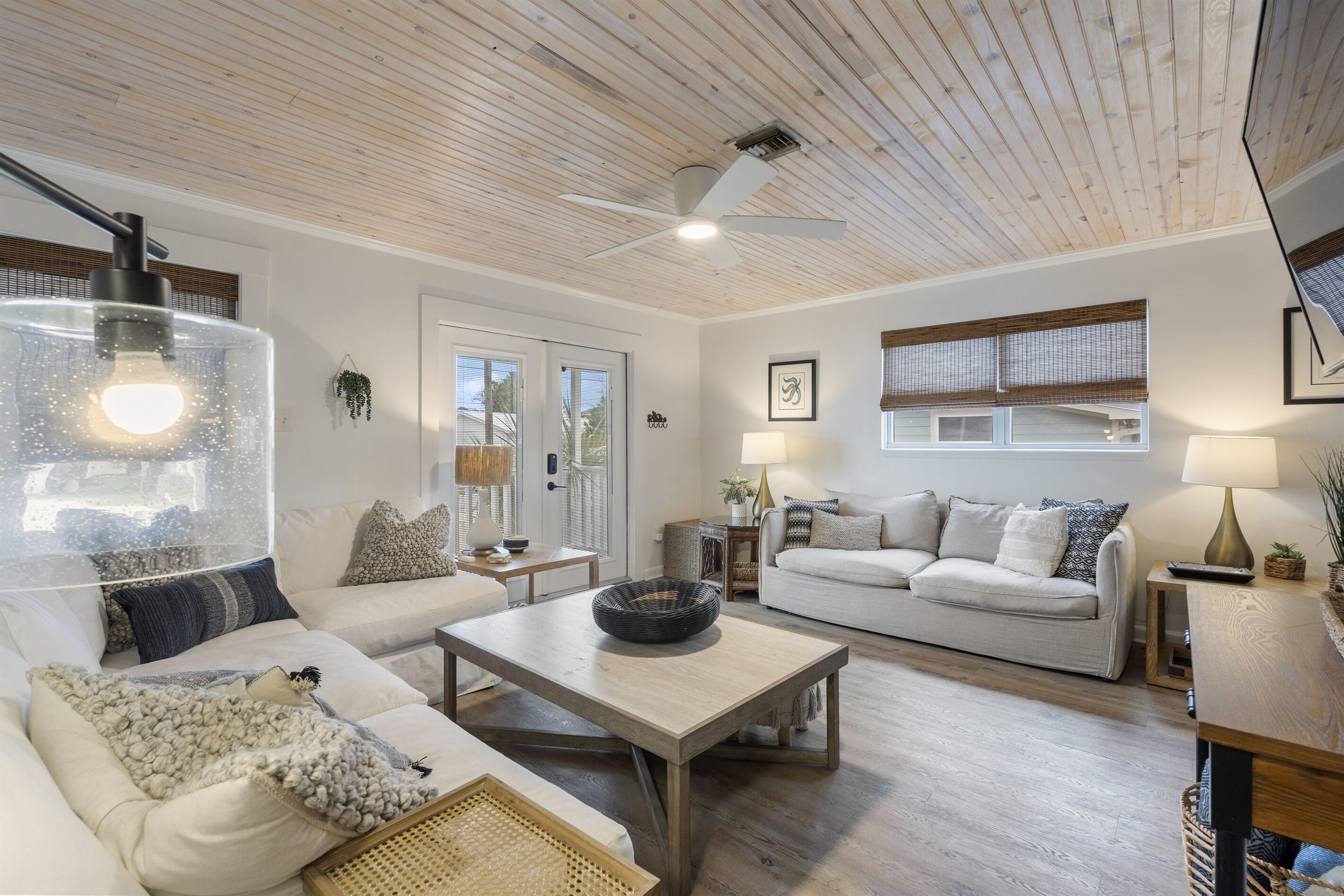 A spacious living room with a wooden ceiling, a ceiling fan, a large window with blinds, a white sofa, a coffee table, and a lamp.