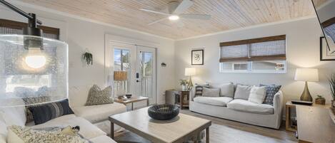A spacious living room with a wooden ceiling, a ceiling fan, a large window with blinds, a white sofa, a coffee table, and a lamp.