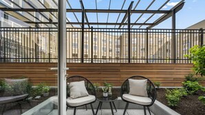 Un patio con dos sillas de mimbre y una mesita pequeña, rodeado por una valla de madera y una barandilla de cristal.