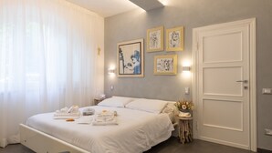 Ein Hotelzimmer mit einem Bett, weißen Vorhängen, einem Nachttisch mit einer Vase und einem an der Wand hängenden Kunstwerk.