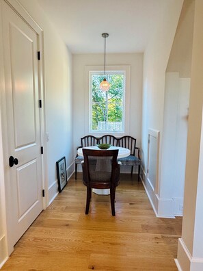 Un pasillo con piso de madera, paredes blancas, una mesa de comedor con sillas y una ventana con vista a árboles.