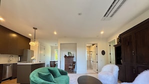 A modern living room with a green sofa, a round coffee table, and a white fluffy chair. There is a kitchen area with dark cabinets and a refrigerator. - Luxury Mountain Retreat with Scenic Balcony Views (Cashiers)