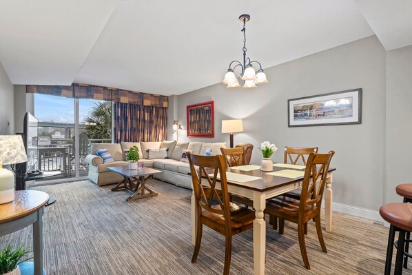 Een moderne woonkamer met een bank, een eettafel en stoelen. Er is een groot raam met gordijnen, een kroonluchter en kunstwerken aan de muren.