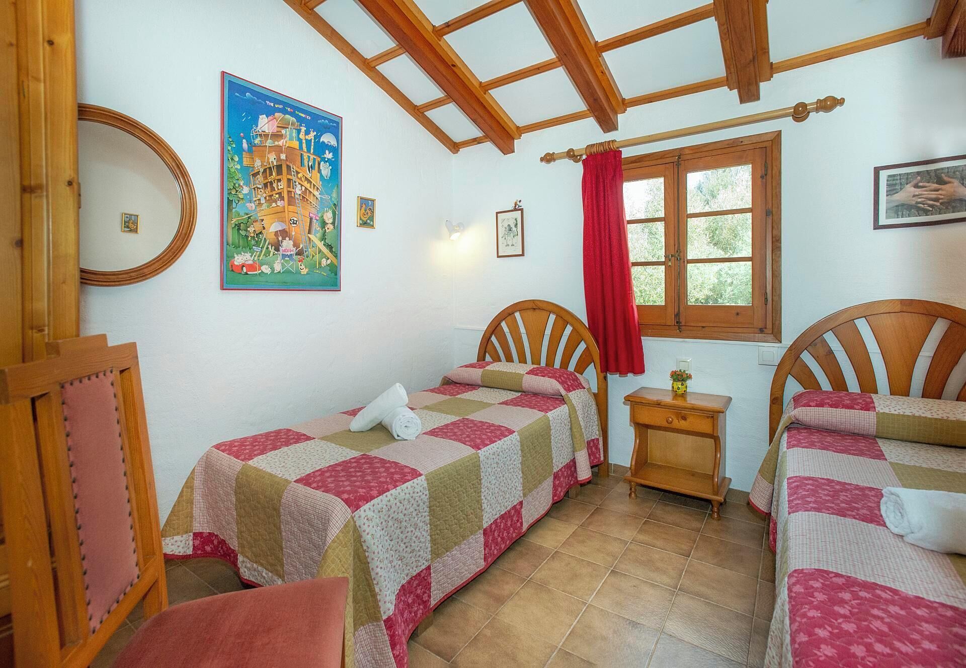 Quarto com duas camas de madeira, uma pequena mesa de cabeceira de madeira, uma janela com cortina vermelha e um quadro na parede.