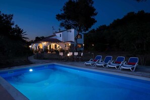 A poolside area with lounge chairs, a dining table, and a house with outdoor lighting. - Sa Mosquera - Four Bedroom Villa, Sleeps 8 (Ciutadella de Menorca)