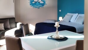 Una habitación de hotel moderna con una mesita de cristal, una vela decorativa, un cuadro azul en la pared, una cama con ropa de cama a rayas y un sofá de color oscuro.