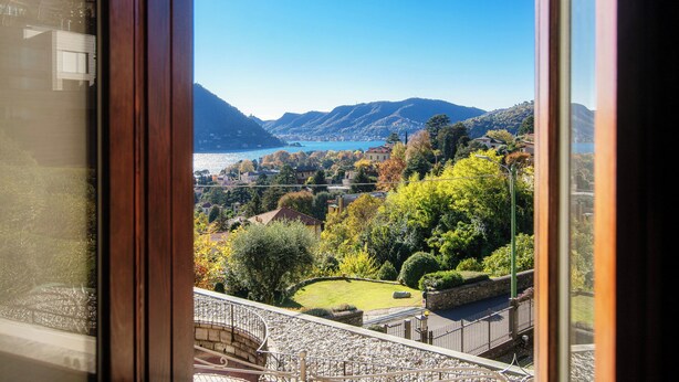 Vista desde una ventana con paisaje montañoso, un lago, un jardín cuidado y un sendero de piedra.