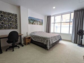 Ein Schlafzimmer mit Bett, Schreibtisch, Stuhl, Standventilator und einem fenster mit Vorhängen.