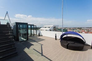 Eine Dachterrasse mit einem gläsernen Anbau, einer Sitzecke mit blauem Baldachin und Blick auf eine Stadtlandschaft.
