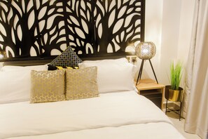 Een net opgemaakt bed met witte lakens en twee decoratieve kussens, een muur met een bladpatroon, een bedlamp en een kleine potplant.