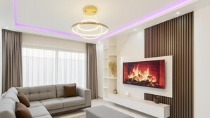 Een moderne woonkamer met een grijze bank, een poef, een flatscreen-televisie en een decoratieve plafondlamp.