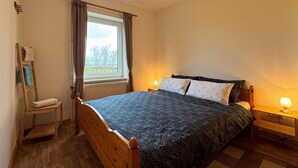 A bedroom with a wooden bed, a nightstand, a chair with towels, a window with curtains, and a view of trees outside.
