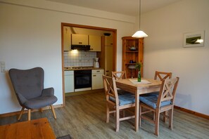A dining area with a wooden table and chairs, a grey armchair, and a kitchen in the background.