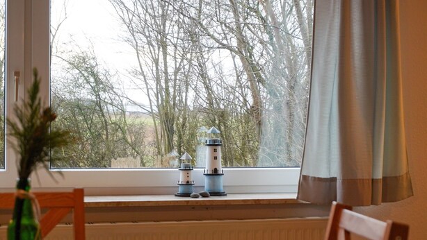 A window with two lighthouses on a sill, a wooden chair, and a table with a vase.