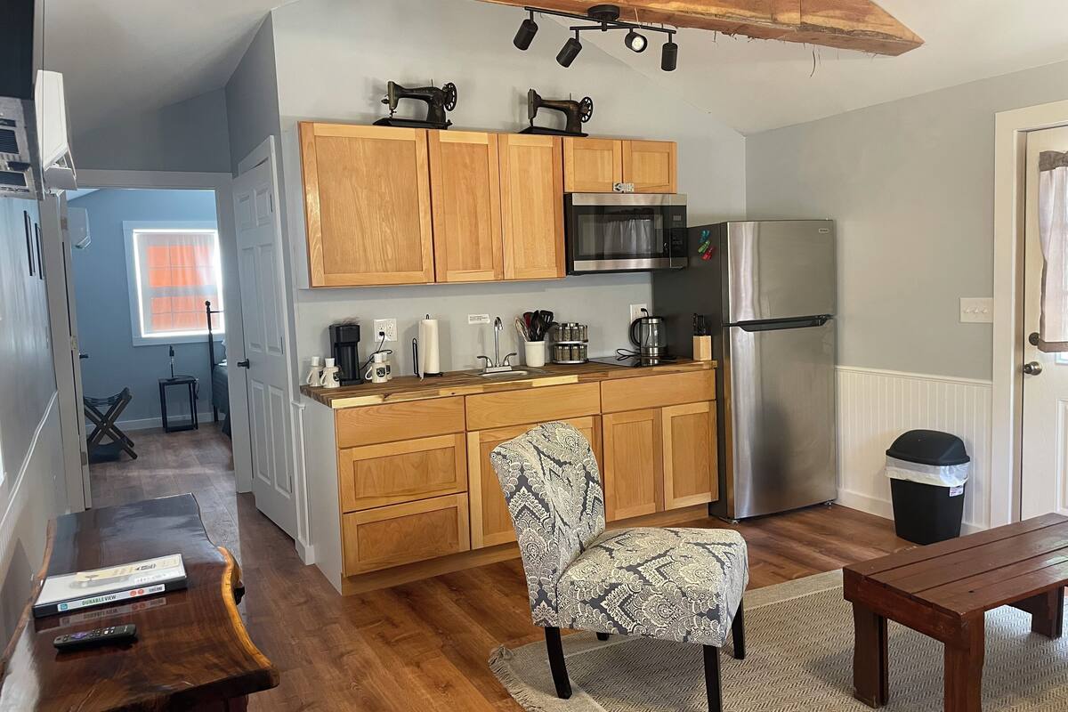 A compact kitchen with wooden cabinets, a stainless steel refrigerator, and a microwave. There's a patterned chair and a wooden table with a remote control on it.