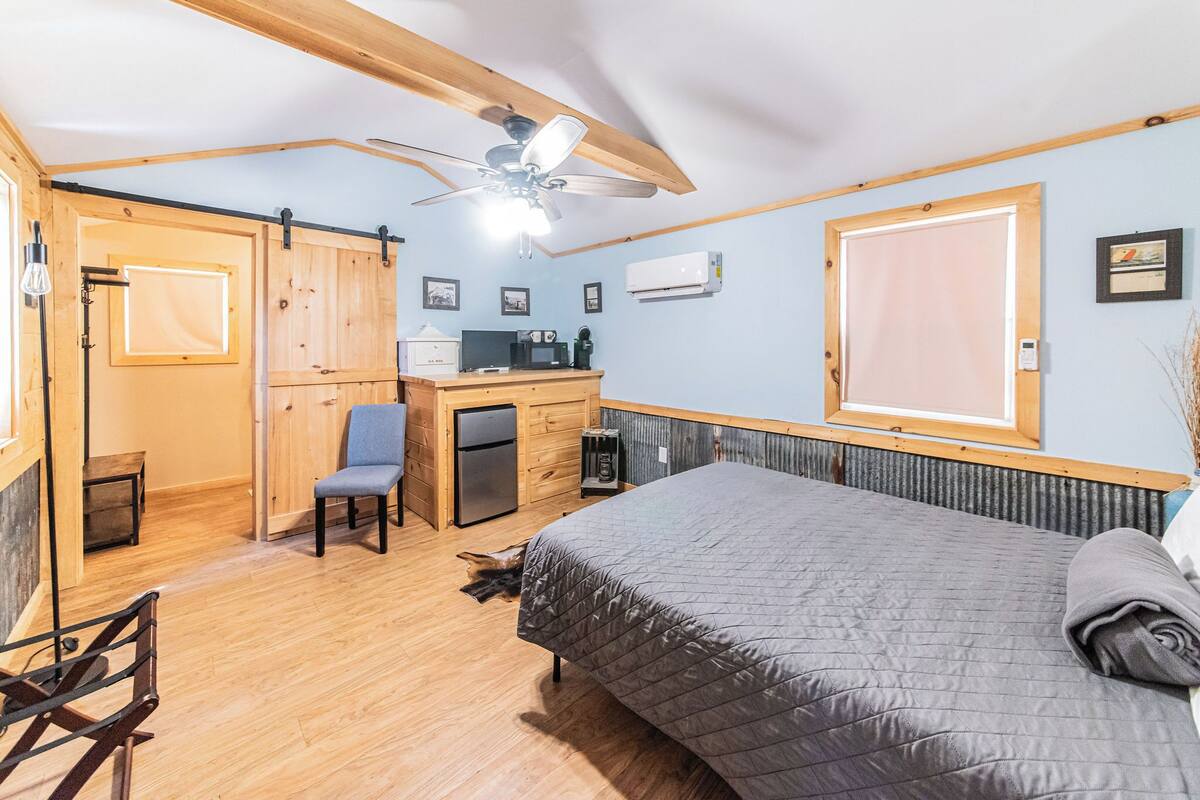 A compact room with a bed, desk, chair, and kitchenette.