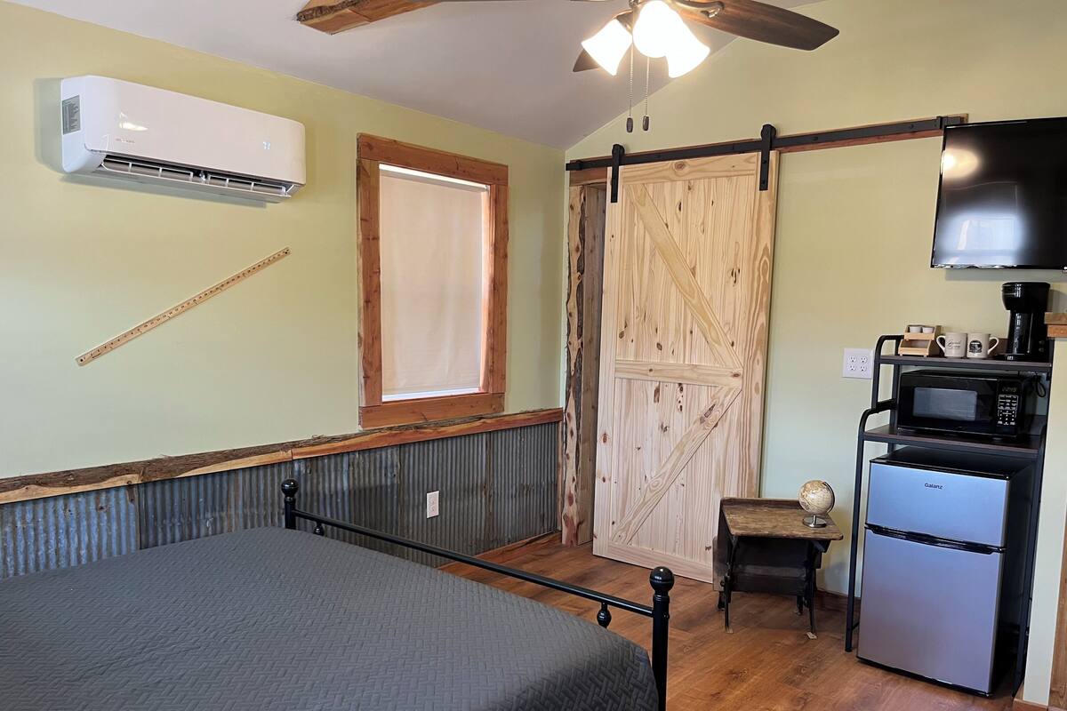 A bedroom with a bed, an air conditioner, a wooden wardrobe, a small table with a globe, a microwave, and a refrigerator.