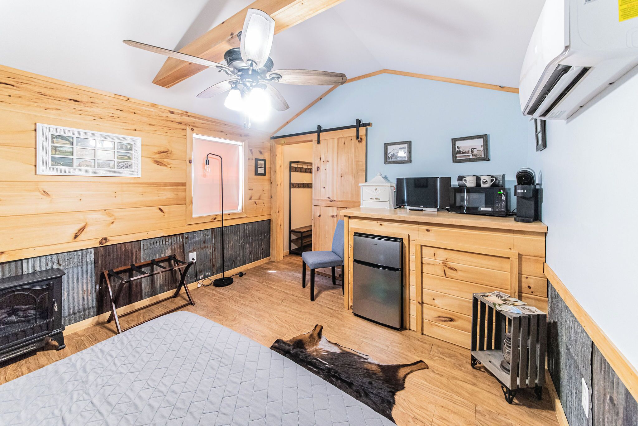 Une chambre confortable, aménagée dans le style d’un chalet, avec un intérieur en bois, un ventilateur de plafond, une petite kitchenette, une cheminée et une grande fenêtre.