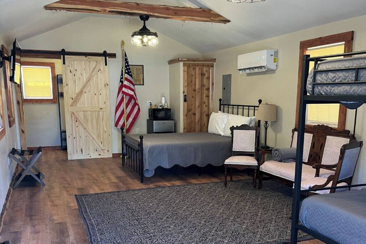 A rustic bedroom with a wooden floor, a bed, a bunk bed, a desk, a chair, a hanging light fixture, and a barn-style door.