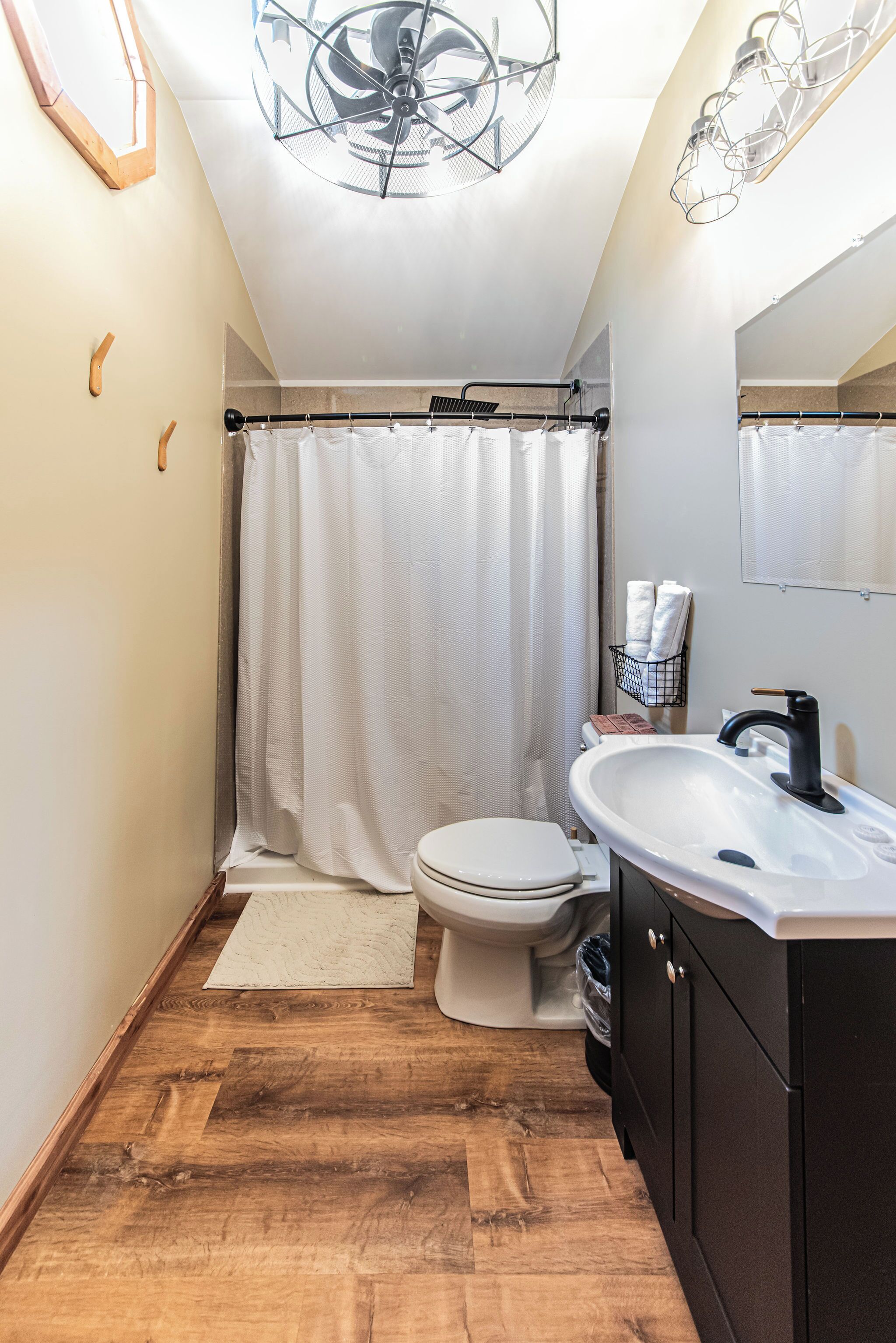 Ein Badezimmer mit Toilette in Weiß, einem schwarzen Waschtisch, einem weißen Waschbecken, einer Duschvorhang und einem Deckenventilator.