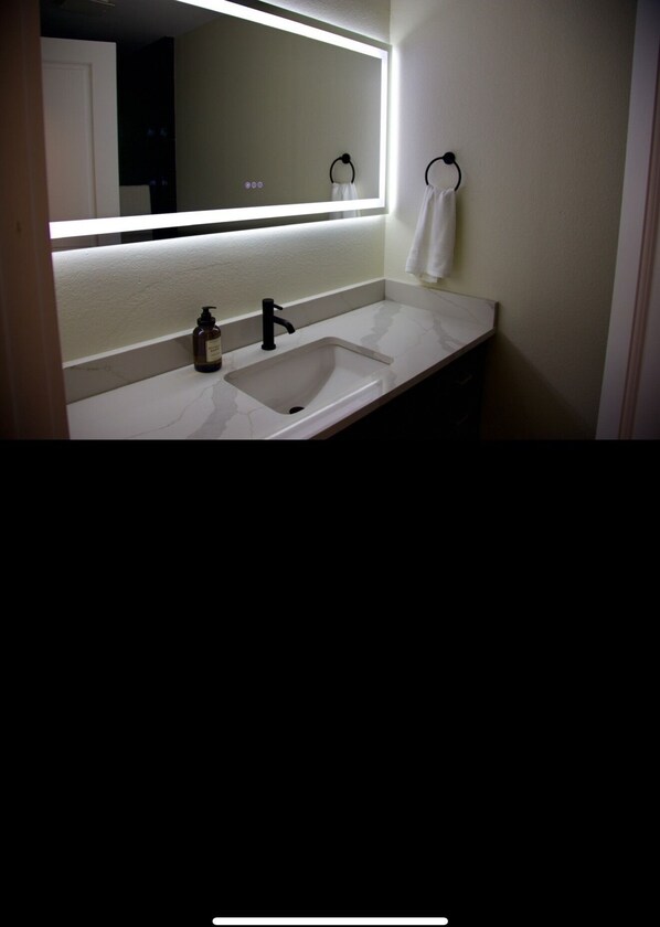 Un baño con lavamanos blanco, grifo negro e iluminación bajo el mueble del espejo.