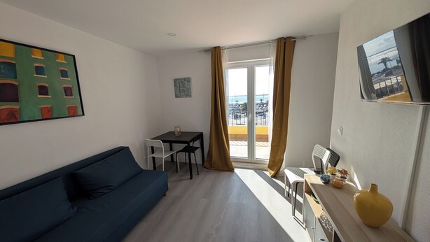 Ein kleiner, heller Raum mit einem blauen Sofa, einem kleinen Tisch mit Stühlen und Meerblick durch große Fenster.