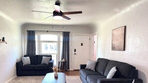 Ein Wohnzimmer mit zwei dunklen Sofas, einem Holztisch als Couchtisch und einem kleinen Beistelltisch. Eine Deckenventilator ist sichtbar, und eine Tür führt in einen anderen Raum.