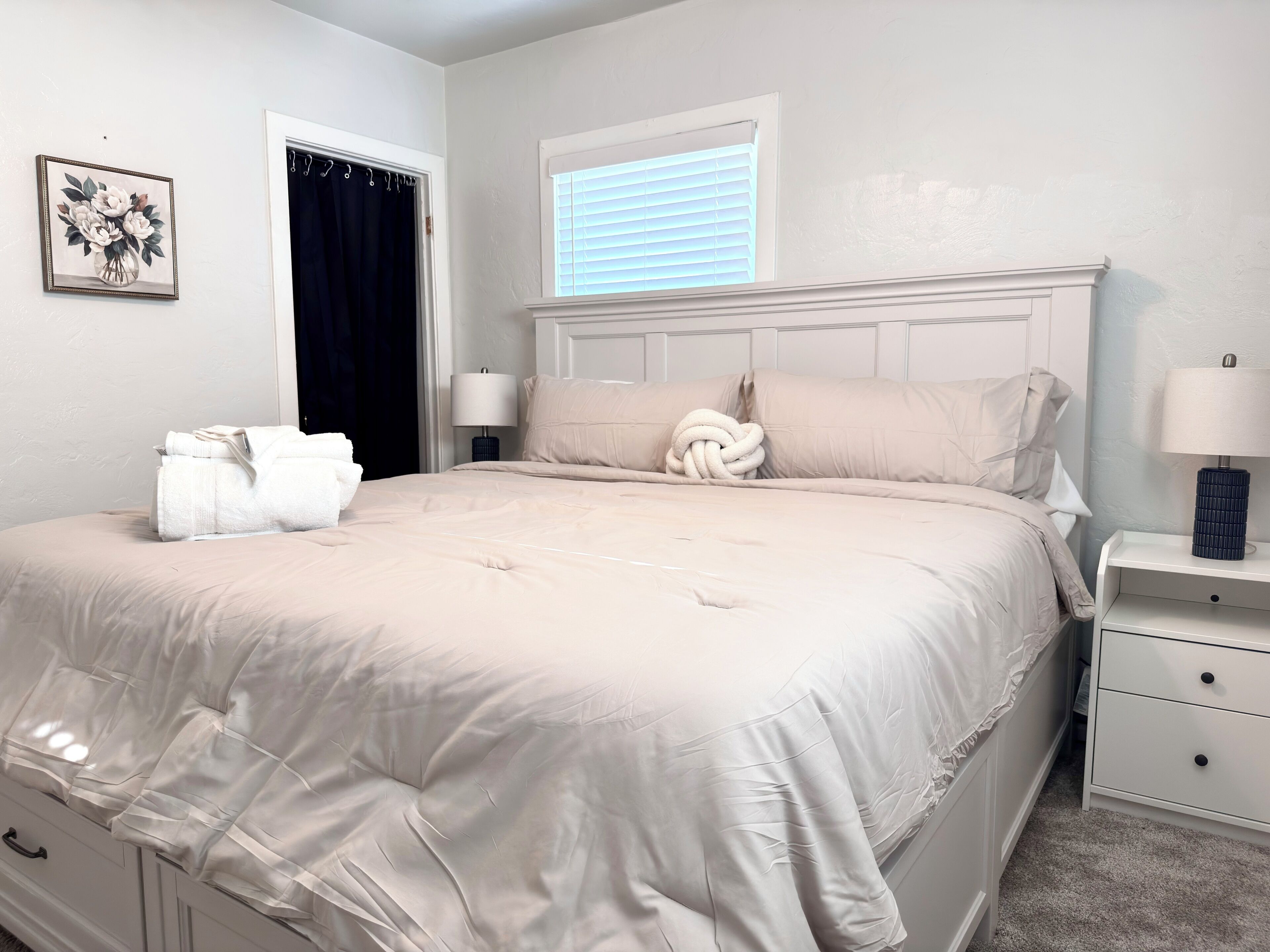 A neatly made bed with a beige comforter, two bedside lamps, a small bedside table with a lamp, a framed floral artwork on the wall, and a window with blinds.