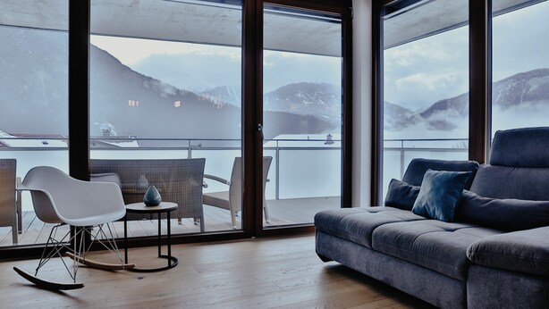 A modern living room with a grey sofa, a rocking chair, and a small table. Large windows offer a view of mountains and a body of water. - Apart Veronika - Exclusive living in Imst (Gemeinde Imst)