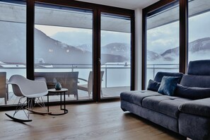 Un salon moderne avec un canapé gris, un fauteuil à bascule et une petite table. De grandes fenêtres offrent une vue sur les montagnes et un plan d’eau.