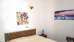 Une chambre à coucher comprenant un lit, une table de chevet, une lampe fixée au mur, une œuvre d’art décorative et une fenêtre donnant sur un paysage.