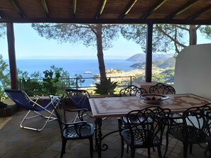 Un área de estar techada al aire libre con una mesa y sillas, con vistas a un paisaje costero y al mar, con colinas lejanas.