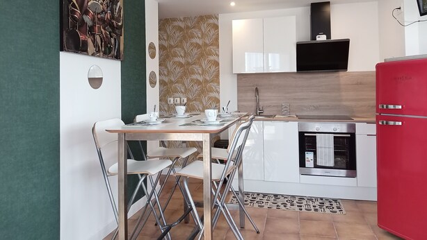 A modern kitchen with white cabinets, a red refrigerator, and a dining area with chairs and a table set for two. - Terra Nova Casa - Free parking 24/24 (Pau)