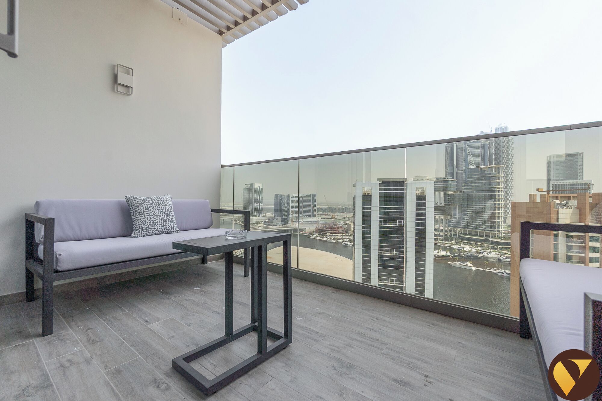 A balcony with a grey sofa, a small table, and a view of a cityscape with high-rise buildings.