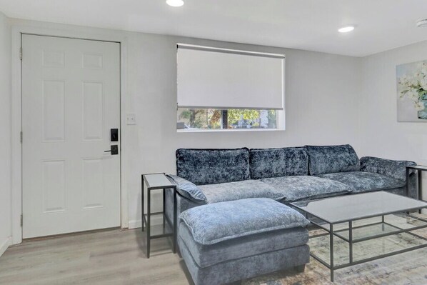 A modern living room with a grey sofa, a glass coffee table, and a window with a white blind.