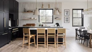 A modern kitchen with black cabinets, a central island with bar stools, pendant lighting, and a dining area with a chandelier. - Lux 5BR South Peak Loon - riverfront w swimming and beach - hot tub - pets ok (Lincoln)