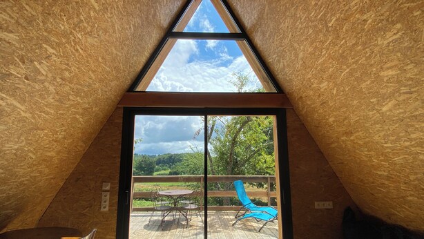 An attic room with a wooden sloped ceiling and a large glass door leading to an outdoor deck with a table, chairs, and a blue chair.