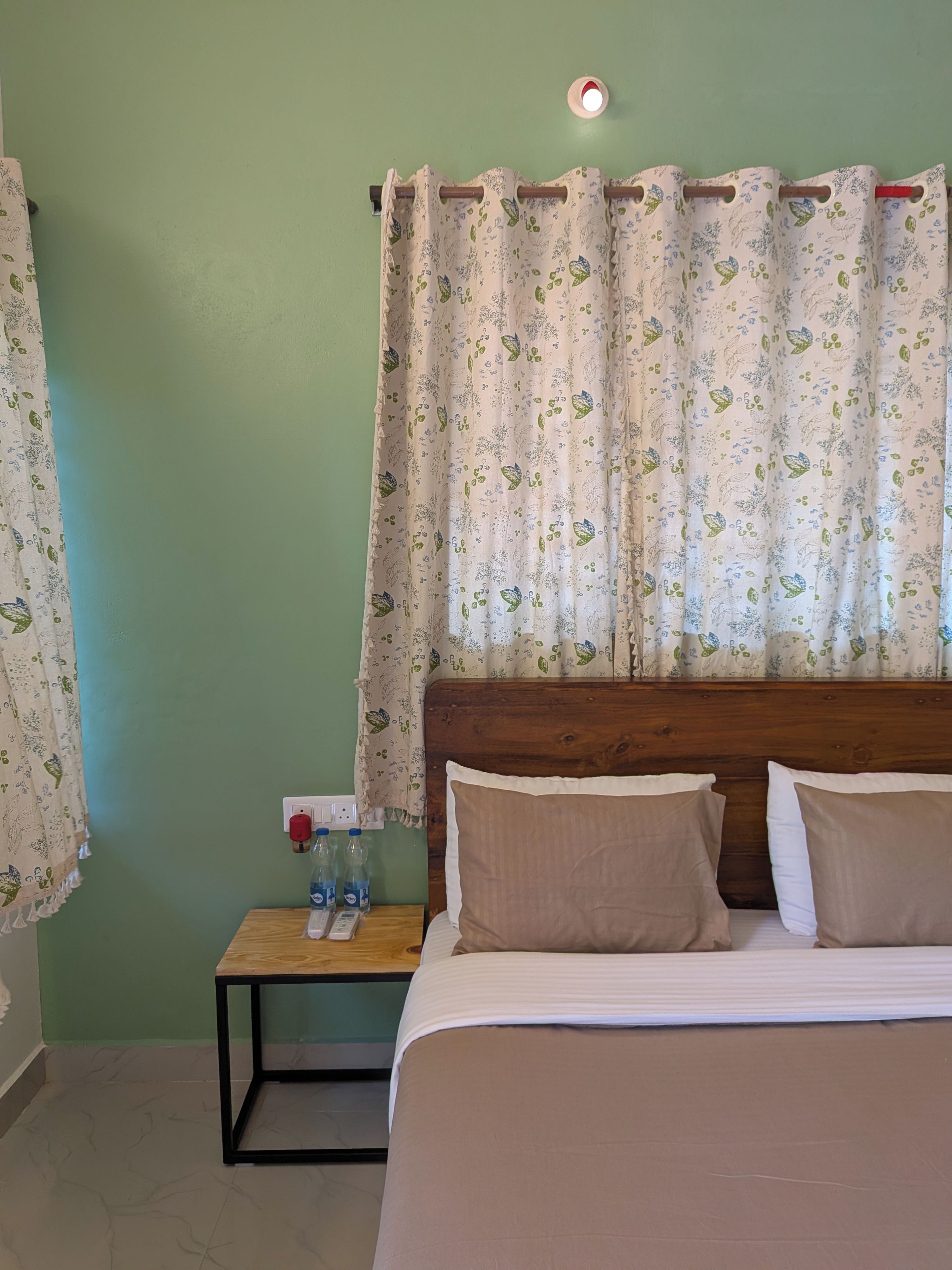 Een slaapkamer met een bed, kussens, een nachtkastje met flesjes en een stopcontact.