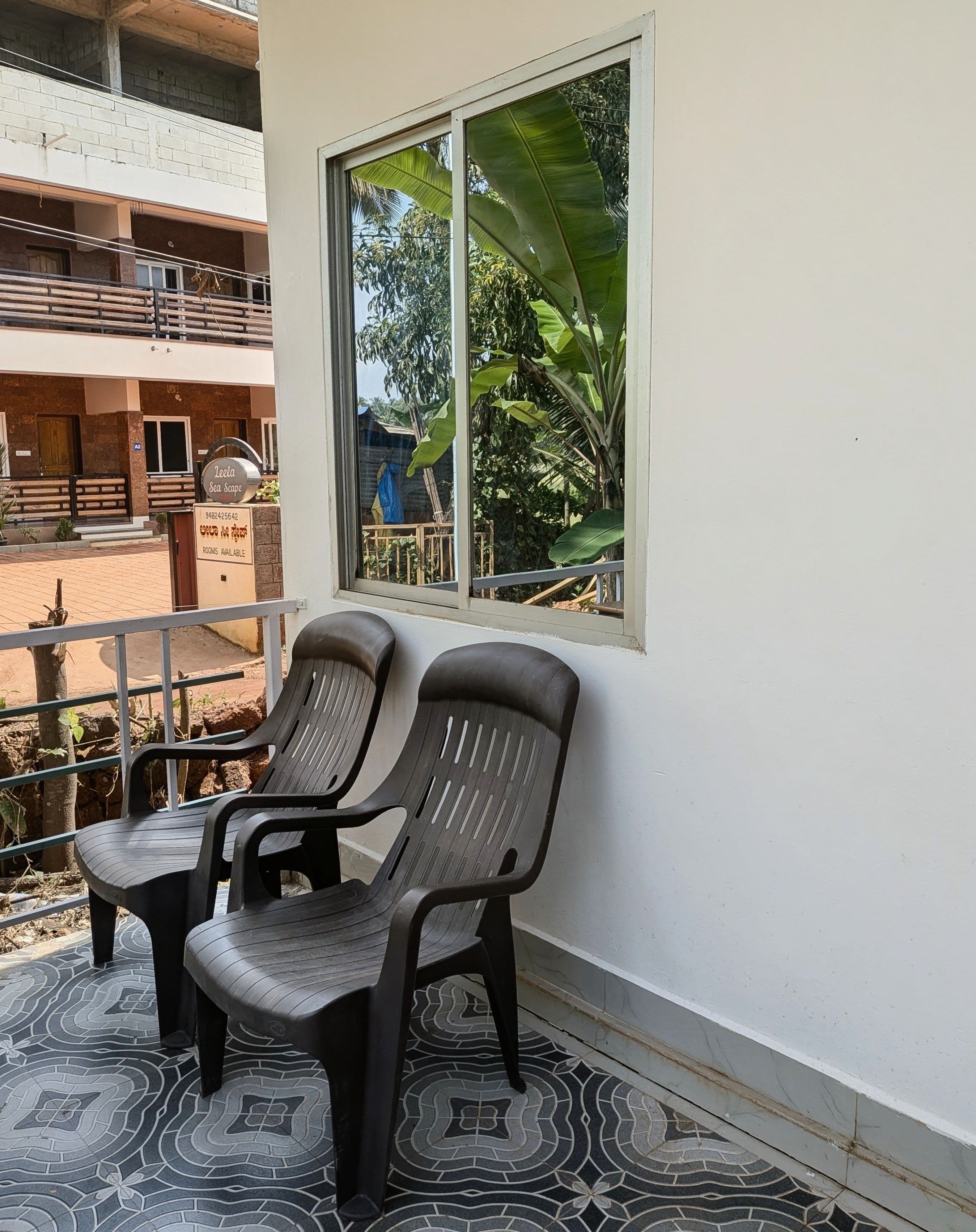 A balcony with two plastic chairs, a window with a view of greenery, and a sign indicating rooms are available.